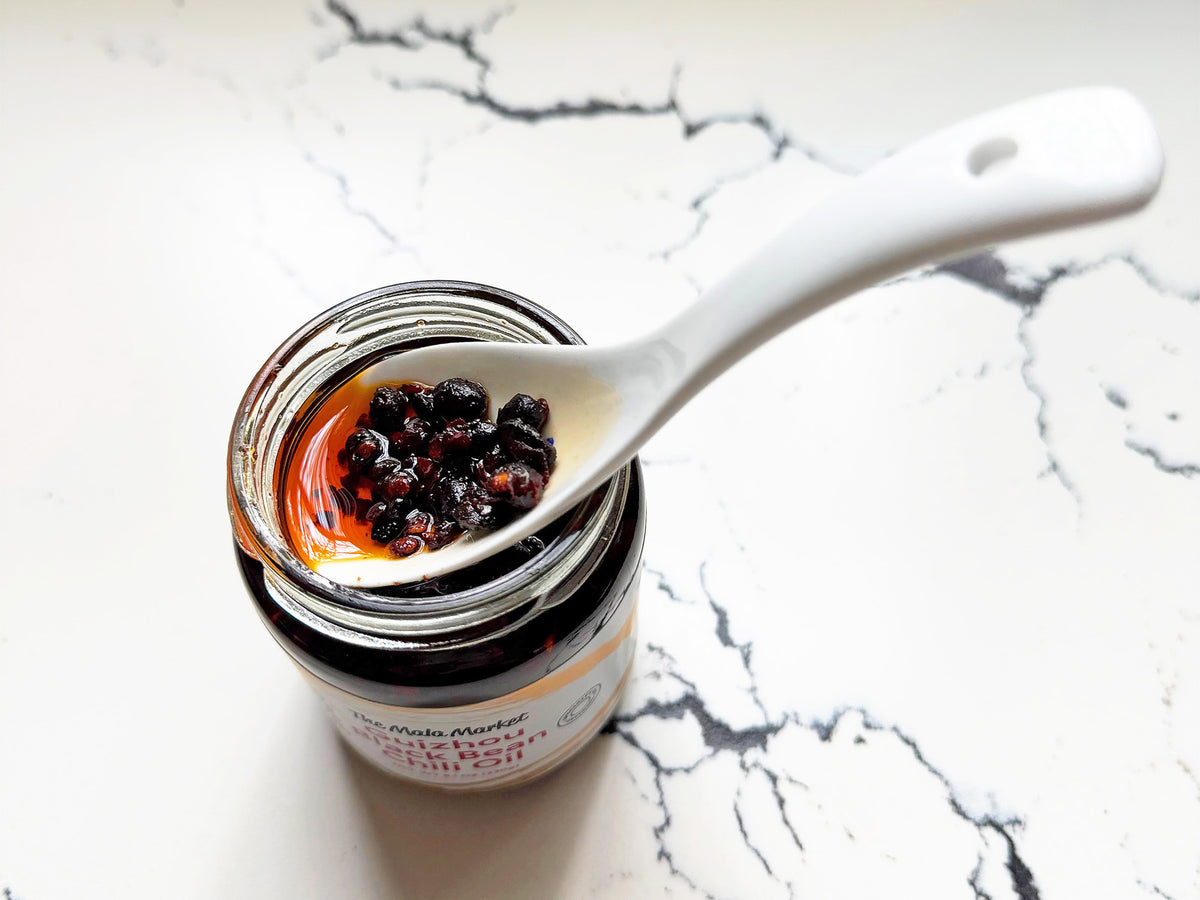 jar of chili oil with the lid off and a white spoon resting on the top holding fermented black beans, chili flakes and oil 