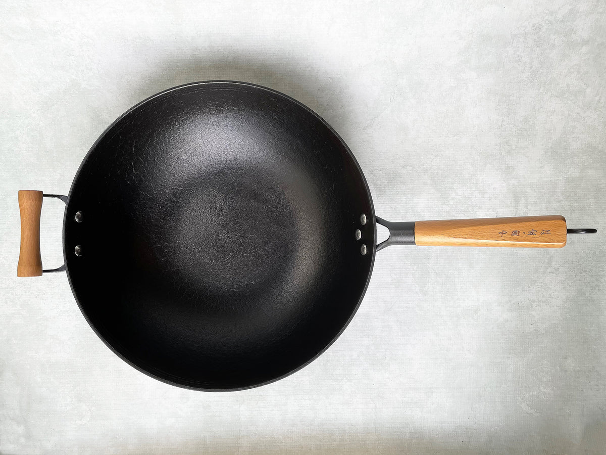 A top view of a black cast iron wok with a flat bottom and a natural oak wood handle