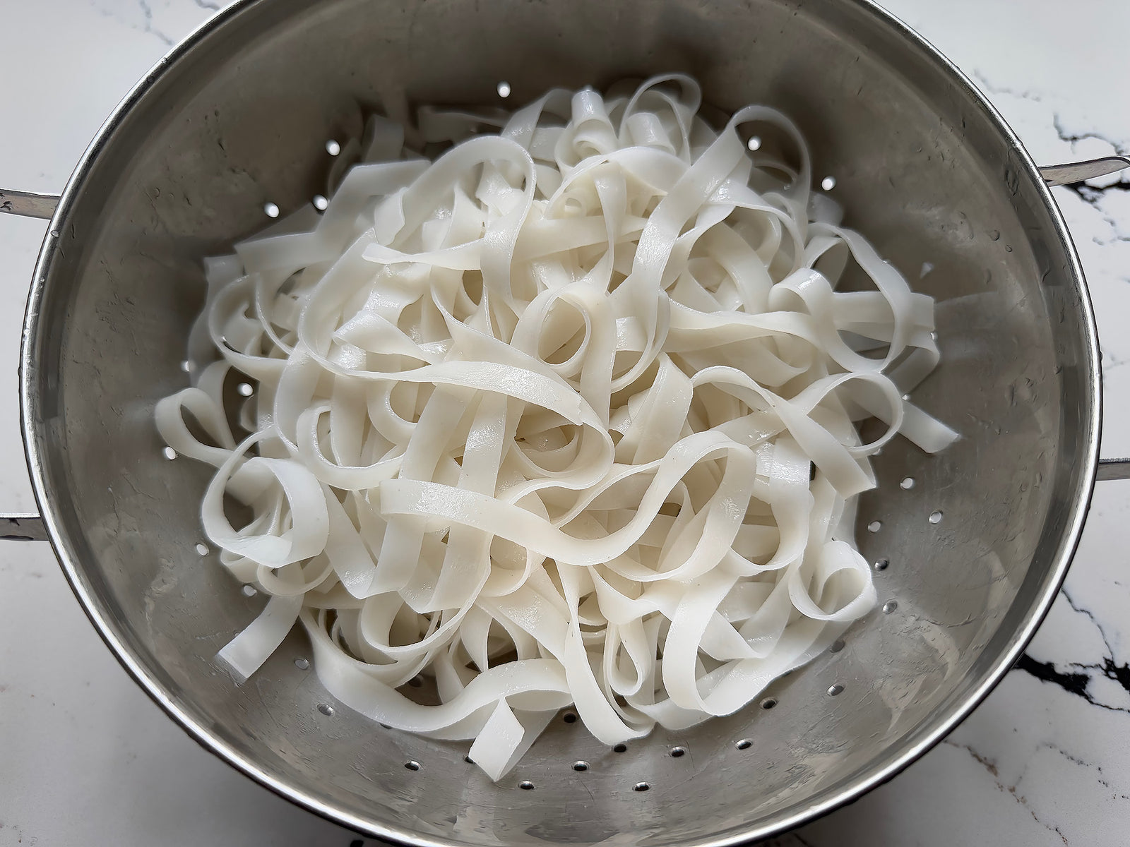 A colander of dried rice noodles that have been soaked and rehydrated