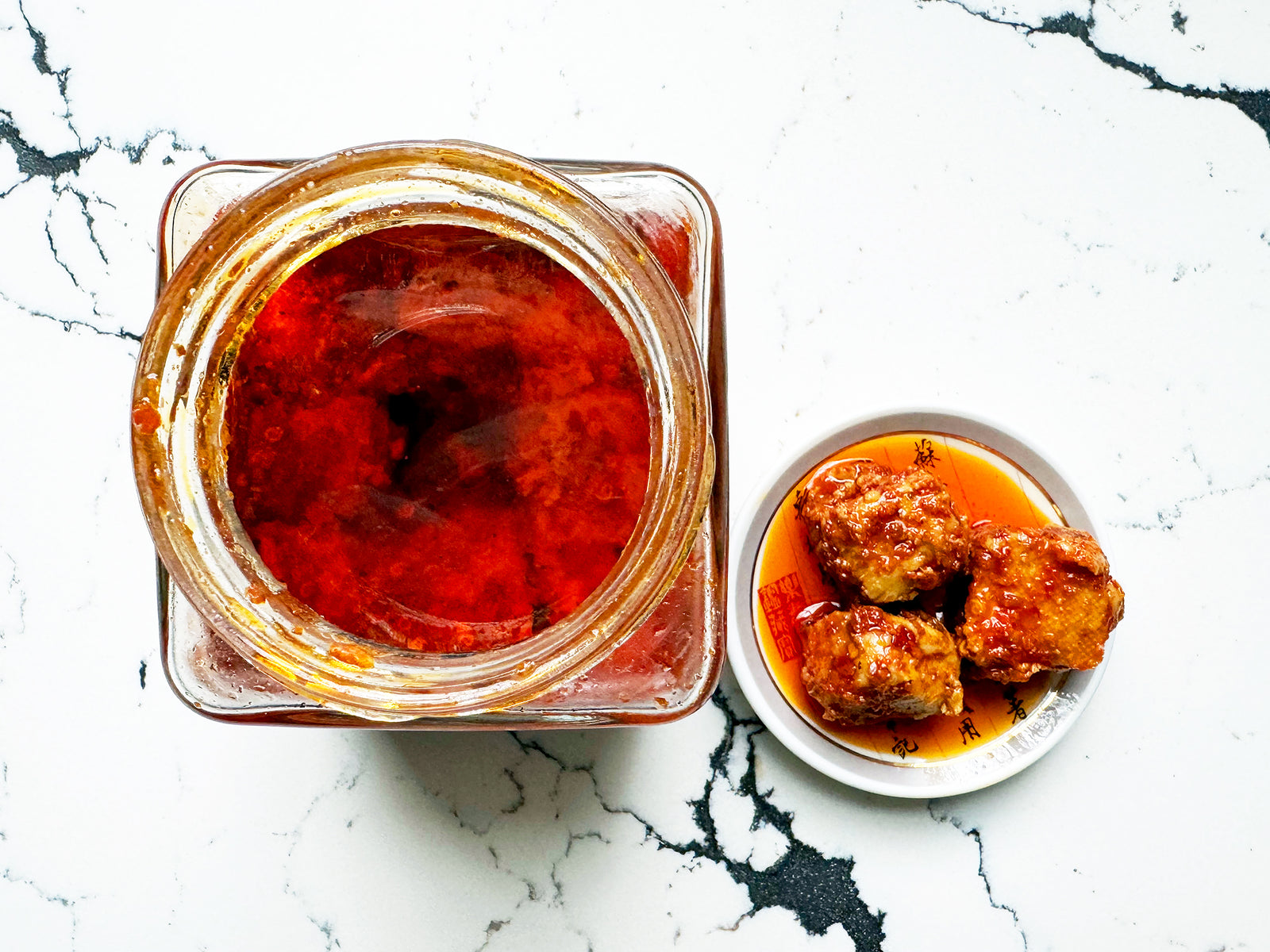 an overhead shot of an open jar showing bright red oil beside a small plate holding three cubes of brownish fermented tofu covered in chili oil 