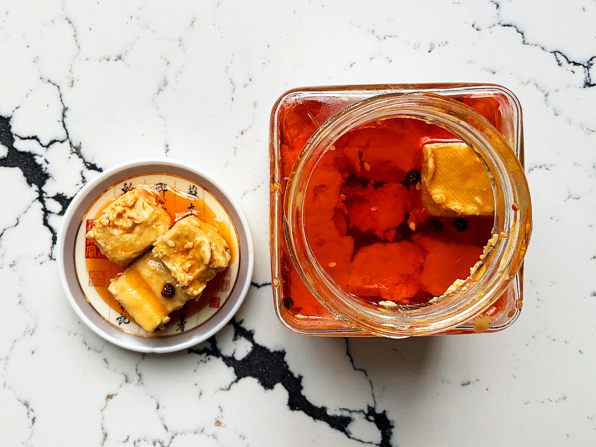 an overhead shot of fermented tofu cubes in a jar covered in clear orange oil and a small plate with three white cubes in a light chili oil