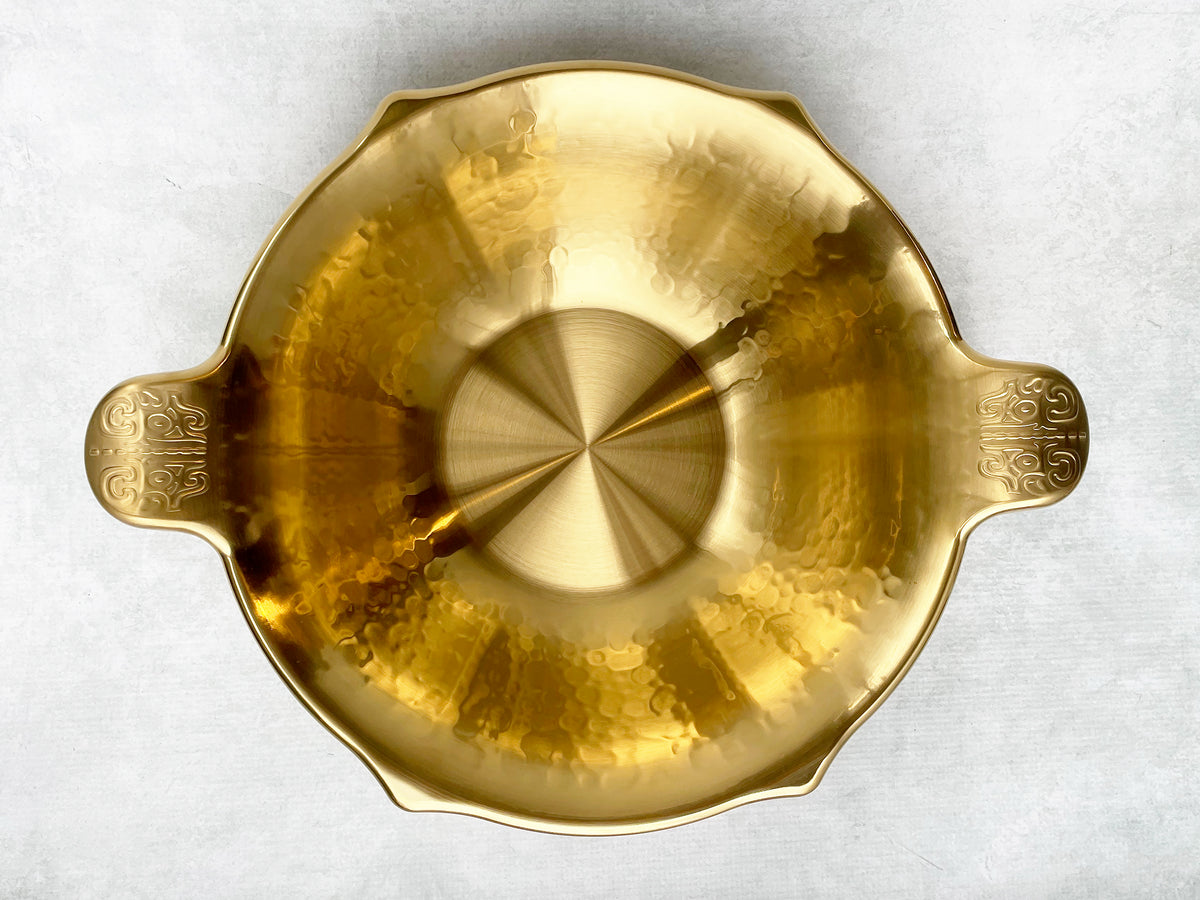 A top view of a gold stainless steel hot pot with scalloped edges and handles with an ancient Asian motif.