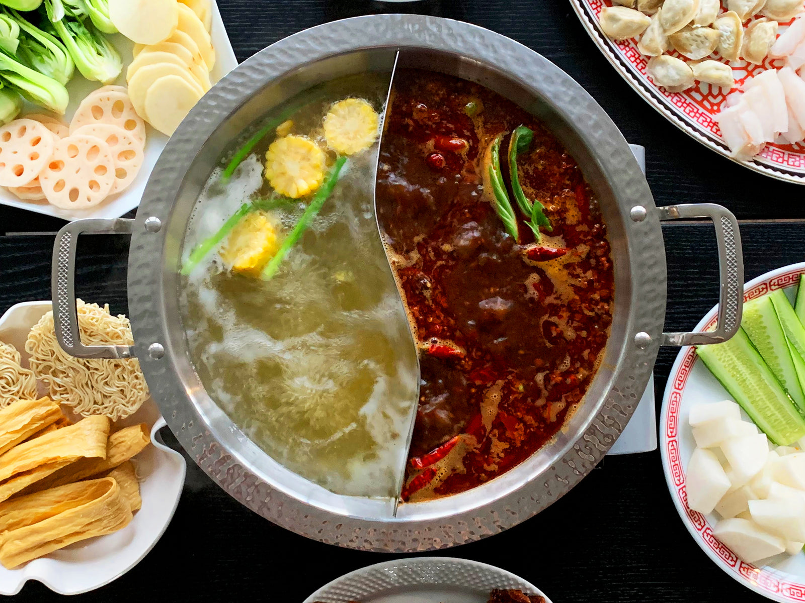 Hot pot with two types of broth surrounded by various ingredients on a dark surface