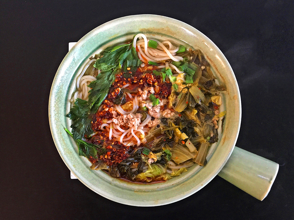 A green clay pot with rice noodle soup topped with pork and pickled greens