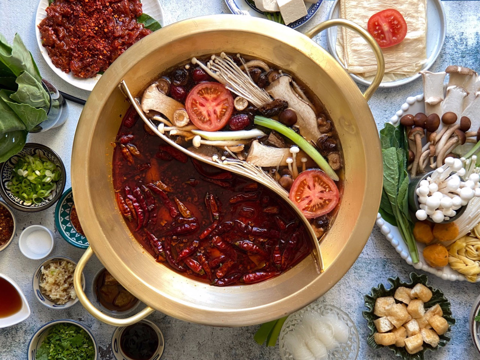 A brass hot pot with mushroom broth and mushrooms on one side and spicy broth with chilies on the other side, with dishes of ingredients alongside