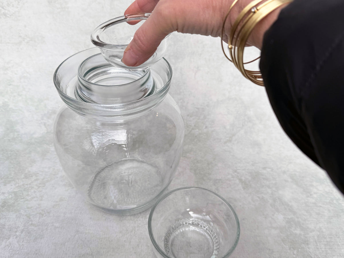 a hand places a glass insert into the top of a glass pickle jar; it is designed to hold down floating vegetables