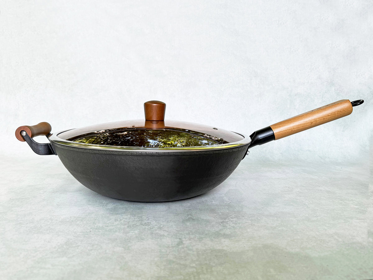 A side view of black cast-iron wok with long wood handle  and short helper handle and glass lid