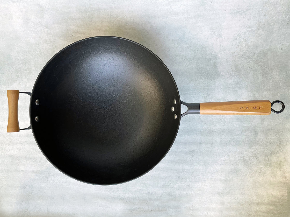 an overhead shot of a round-bottom cast-iron wok with a long wood handle and a short wood helper handle