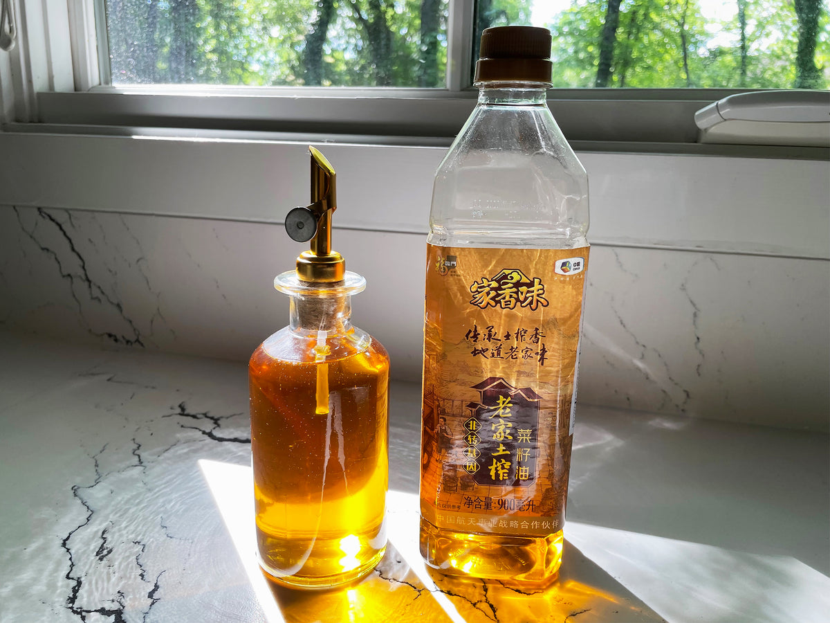 a half-empty plastic bottle of golden caiziyou and a decorative glass bottle full of the roasted rapeseed oil with a pour spout, reflecting sunlight from a window