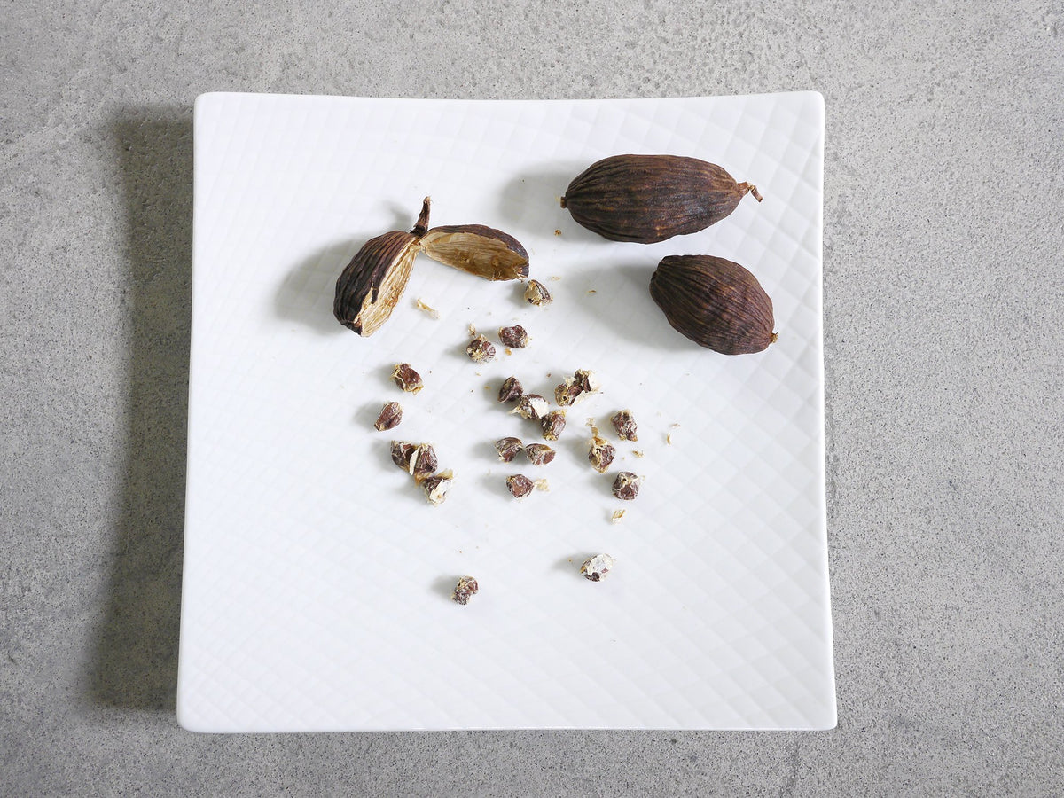 two whole black cardamom pods on a white plate and another pod split open to reveal many small seeds