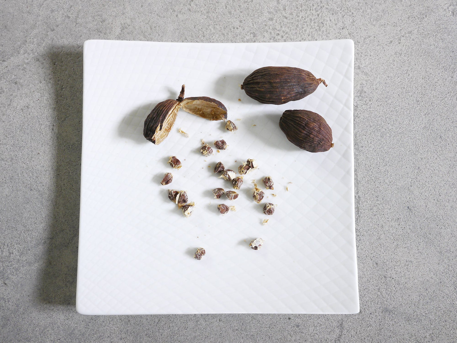 two whole black cardamom pods on a white plate and another pod split open to reveal many small seeds