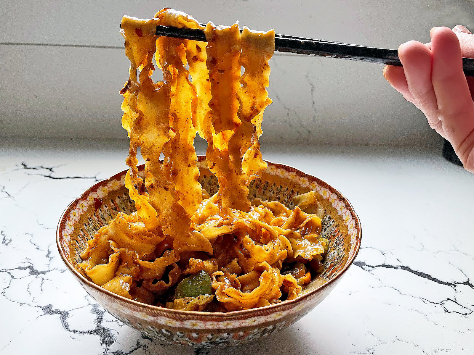 knife-cut noodles in a red chili meat sauce being lifted from a bowl by chopsticks 