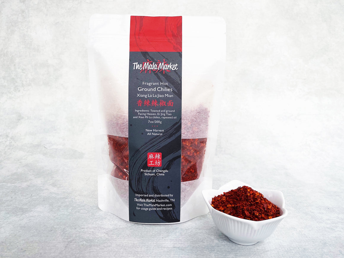  a white kraft bag of red ground chilies and a small white bowl next to it displaying the same ground chilies