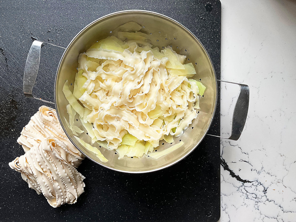 a metal colander of cooked knife-cut noodles on top of cooked cabbage and a couple uncooked bundles of noodles beside it